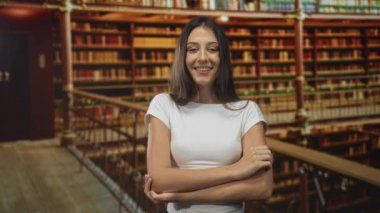 Elleri çenesinde gülümseyen ve kütüphane kitaplarıyla kollarını kavuşturan bir kadın..