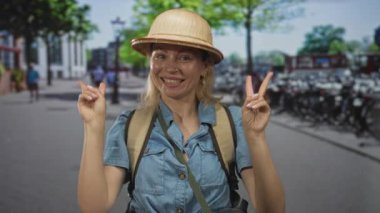 Sırt çantalı kadın güneşli şehir caddesinde gülümserken iki eliyle barış işareti gösteriyor. Gezgin şehvet neşesi..