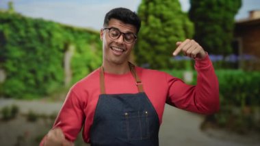 Gözlüklü ve önlüklü genç adam gülümsüyor ve yeşillik ve patikalarla çevrili bir açık hava parkına işaret ediyor..