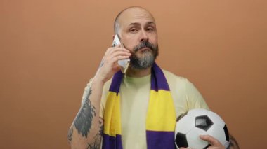 Telefondan konuşan sakallı bir adam futbol topunu tutuyor ve kahverengi arka planda eşarp takıyor.