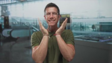 Havalimanı terminalinde yürüyen merdiven ve bagaj atlıkarıncasında avuçlarını çırpan, gözlüklü ve yeşil tişörtlü adam; sevinç kutlamaları.