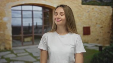 Açık havada, gözünün yanındaki parmağıyla el kol hareketi yapan, gözleri kapalı taş bir duvara poz veren, rahat bir gülümseme takınan, gözlem ve farkındalık hissi veren genç bir kadın..