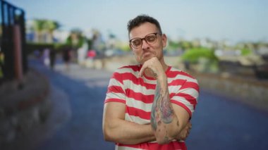 Dövmeli ve gözlüklü genç bir adam, parlak bir şehir sokağında kenar çizgili bir gömlek ve bulanık bir şehir arka planıyla güvenle dururken iyi bir jest yapıyor..