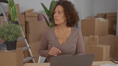 Kadın, dizüstü bilgisayarın yanında istiflenmiş karton kutulara ve yeşil saksı bitkisine işaret ediyor..
