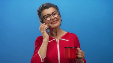Gri saçlı ve gözlüklü yaşlı kadın telefonda konuşurken gülümsüyor. Kırmızı kahve fincanını mavi arka planda tutuyor..