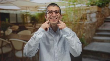 Restoranın terasında çizgili tişörtle gülümseyen bir adam, parlak, doğal bir ortamda mutluluğu sergiliyor, taş yol boyunca görünen yeşillik..