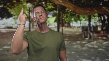 Yeşil gömlekli bir adam bir girişin yakınındaki benekli ağaç gölgesinin altında gözlük takarken işaret parmağını işaret ediyor..