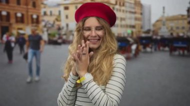 Kadın, plazada bir kalp şekli oluşturmak için parmaklarını kullanır. Sarışın, genç, gülümseyen bir denekle. Bereli, gözlüklü ve çizgili bir bluzla..