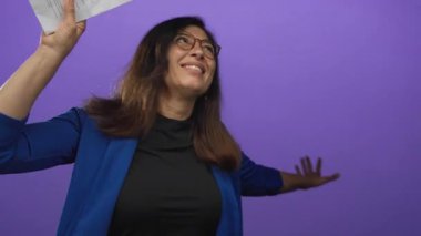 Orta yaşlı, gözlüklü bir kadın sarı pano tutuyor ve stüdyoda mor duvara uzanmış el hareketleriyle sevinç ifadesi iletişimi kuruyor..