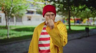 Sarı ceketli genç adam parkta jest yapıyor. Arka planda yeşil ağaçlar var.