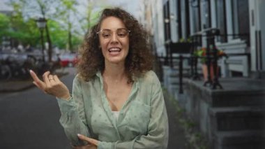 Elleriyle hareket eden gözlük takan ve şehir sokaklarında gülümseyen bir kadın bina merdivenlerine yaslanırken neşeli bir ifade..