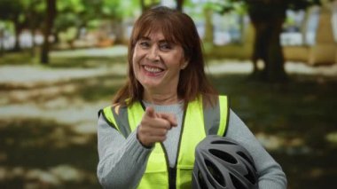 Latin kökenli son sınıf öğrencisi bir kadın parkta kameraya bakarken, yansıtıcı bir yelek giyip, bisiklet kaskı tutarken, açık hava aktivitelerinde güvenliği somutlaştırırken gülümsüyor..