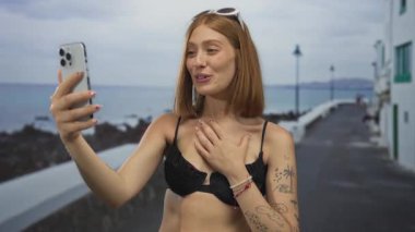 Göğüs göğüse gezen genç sarışın kadın deniz kenarındaki bir gezinti alanında akıllı telefondan video görüşmesi yapıyor..