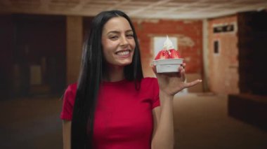 Kapalı bir inşaat alanında elinde tatlıyla gülümseyen bir kadın parlak kırmızı gömlekli, uzun siyah saçlı ve bitmemiş arka planda neşeli bir ifade sergiliyor..