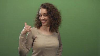 Stüdyoda yumrukla gülümseyen, yeşil ekranlı bir kadın, elleri kalçasında görünür, kendine güveni tam..