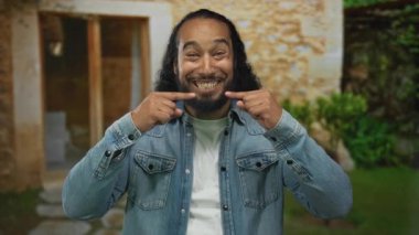 Adam dışarıdaki bir binanın girişinde, kot ceketle gülümseyen dişlerini işaret eder..