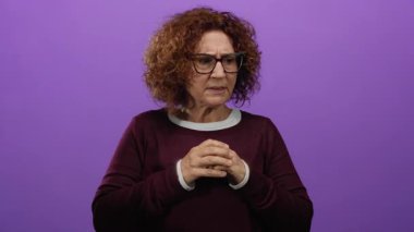 Orta yaşlı, gözlüklü bir İspanyol kadın mor bir duvarın önünde endişeli görünüyor. El hareketleriyle duygularını ifade ediyor..