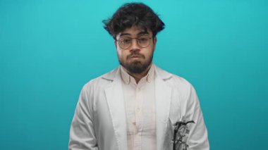 Laboratuvar önlüklü, gözlüklü ve sakallı genç adam mavi bir arka plana karşı duruyor. Profesyonel bir tıbbi ortamda İspanyol bir doktor ya da bilim adamını canlandırıyor..