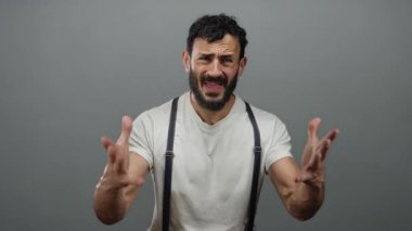 Sakallı İspanyol bir adam beyaz gömlekli, pantolon askısı takmış gri bir arka planda hayal kırıklığı yaşıyor..