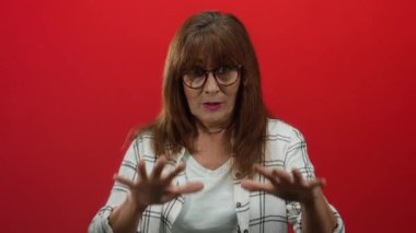 Kıdemli İspanyol kadın, canlı kırmızı bir duvara karşı sakince el kol hareketi yapıyor. Gözlüklü ve ekose gömlekli, orta boylu saçlı, kapalı alanda izole edilmiş, düşünceli bir sahne yaratıyor..