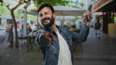 Sakallı İspanyol bir adam araba anahtarlarını güvenli bir şekilde bir sokak ortamında tutuyor, kot ceket giyiyor ve canlı bir şehir ortamında ağaçların altında gülümsüyor..