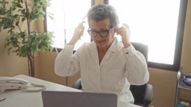 Klinikte elinde steteskop, yanında ofis ekipmanları ve bir bitkiyle gülümseyen yaşlı kadın doktor..