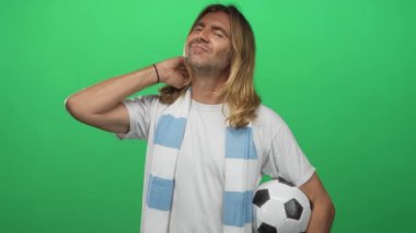 Kaba sarışın adam saçlarına dokunuyor ve futbol topunu canlı yeşil stüdyo ortamında tutuyor..