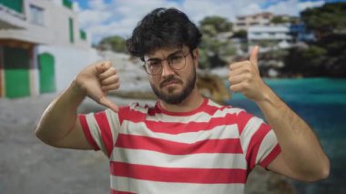 Sakallı, çizgili tişört giyen genç adam deniz kıyısında, bina ve okyanusun arka planında, açık havada karışık duygular olduğunu gösteren işaretler yapıyor..