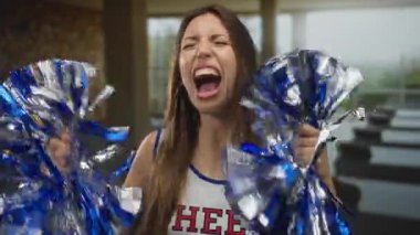 Uzun kahverengi saçlı beyaz ponpon kadın metalik mavi ponponu sallıyor ve bina içinde ağzını açıyor..