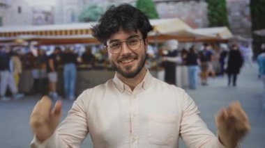 Sakallı, yumruklarıyla kutlama yapan genç bir İspanyol adam gün içinde insan ve tezgahtarlarla dolu bir açık pazar caddesinde duruyor..