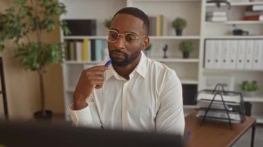 Gözlüklü ve beyaz gömlekli bir adam ofis masasında çenesine mavi bir kalem tutuyordu. Yanında da raflar ve bitkiler vardı..