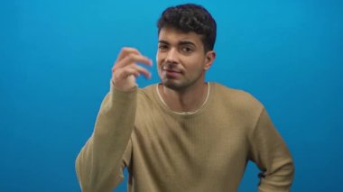 Adam sağlam bir arkaplanda omuz silkip elini kaldırıyor. Mavi şaşkın süveter ve erkek ifadesiyle Latin kökenli genç bir şahıs öne çıkıyor..