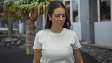 Modern bina cephesi ve avlu girişinde elleri başının üzerinde olan beyaz tişörtlü genç esmer kadın; stres reaksiyonu.