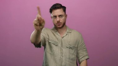 Sakallı genç adam, izole edilmiş pembe bir arka plana karşı onaylanmamış bir jest yapıp bej gömlekli, iddialı bir ifade sergiliyor..