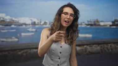Gözlük takan genç İspanyol kadın ve açık gri yelek parmağıyla deniz kenarındaki caddeyi işaret ediyor..