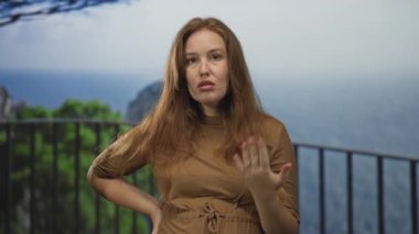 Kızıl saçlı kadın, ağaçlara ve denize tepeden bakan korkulukların yanında dururken el ele tutuşuyor..