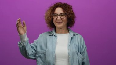 İspanyol orta yaşlı, kot gömlekli, kıvırcık saçlı kadın izole edilmiş pembe arka plan duvarına uzanmış, sıcaklığı ve arkadaşlığı yayıyor..