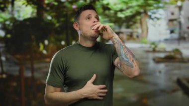 Şehir merkezinde hapşıran, dövmeleri görünen genç adam, bulanık sokak arka planının ortasında yeşil gömleğin altında rahatsızlığını ifade ediyor..