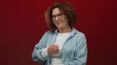 Hispanic woman with glasses embracing herself stands against a vivid red wall, exuding warmth and self-love.