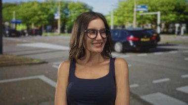 Sıradan donanma tişörtlü ve büyük gözlüklü bir kadın güneşli şehir kaldırımında bulanık arabalarıyla gülümsüyor..