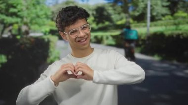 Gözlüklü genç İspanyol adam, yeşil ağaçlar ve güneş ışığıyla kaplı caddede kamerayı işaret ediyor..