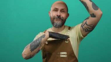 Sakallı, yeşil bir duvara karşı kahverengi tulumlu, dövmeli ve siyah eldiven giyen kendine güvenen beyaz bir adam..