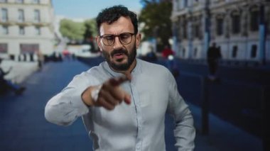 Sakallı ve gözlüklü İspanyol bir adam kalabalık bir sokakta şehir ortamında ciddi bir ifadeyle dikiliyor..