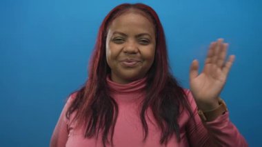 Afro-Amerikalı bir kadın stüdyoda pembe bir balıkçı yaka giyip el sallıyor ve şık bir şekilde gülümsüyor..