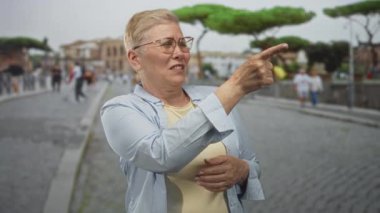 Kadın, kaldırım taşı caddesini işaret ediyor. Gözlük takıyor ve hafif bir gömlek giyiyor. Uzak yayaların arasında sağ tarafa işaret ediyor..