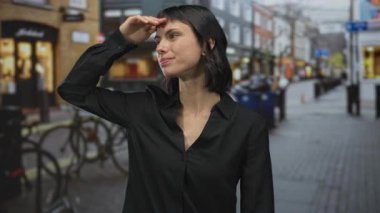 Siyah tişörtlü genç İspanyol kadın yoğun caddede alnında elleriyle gözlerini koruyor; merak.