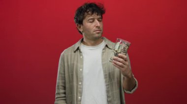 Hispanik adamın elinde dolarla dolu bir kumbara var. Kırmızı arka planda hareketli bir el hareketi yaparak tasarruf ve finansal ihtiyat öneriyor..