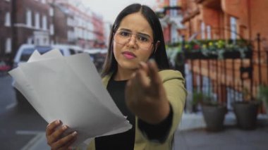 Şehir merkezinde elinde gözlüklü ve ceketli bir kadın, açık şehir ortamında ifadesini belirterek soruşturma ya da çatışmaya işaret ediyor..