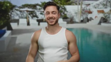 Otelin bilardo salonundaki kameraya gülümseyen sakallı genç adam, beyaz atlet giyiyor, el hareketi yapıyor, güneşli gökyüzünün altında açık hava otelinin tadını çıkarıyor..