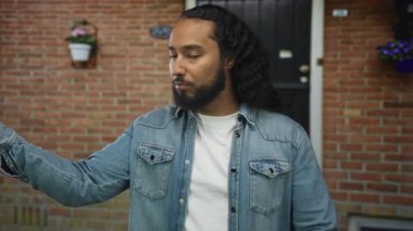 İşaret parmağıyla evin dışındaki bir binanın girişine işaret eden adam, kot ceket ve beyaz tişört giyiyor, sakalı görünüyor; iddialı uyarı.